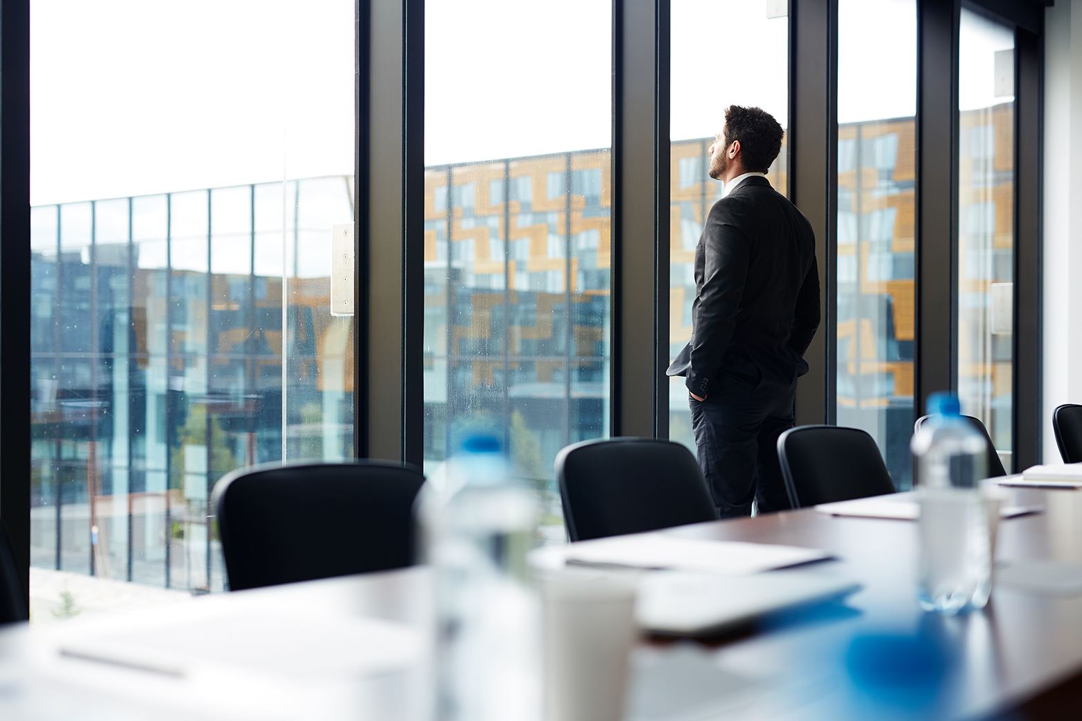 Back view portrait of successful businessman standing by window and looking outside with his hands in pockets, copy space